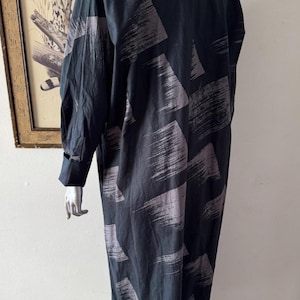 K&ouml;nnte beinhalten: Ein langes, schwarzes Kleid mit einem grauen Pinselstrichmuster. Das Kleid hat lange &Auml;rmel mit gekn&ouml;pften Manschetten. Das Kleidungsst&uuml;ck wird auf einer Schaufensterpuppe pr&auml;sentiert. Der Hintergrund umfasst ein gerahmtes Bild eines Leoparden und eine dekorative Feder.