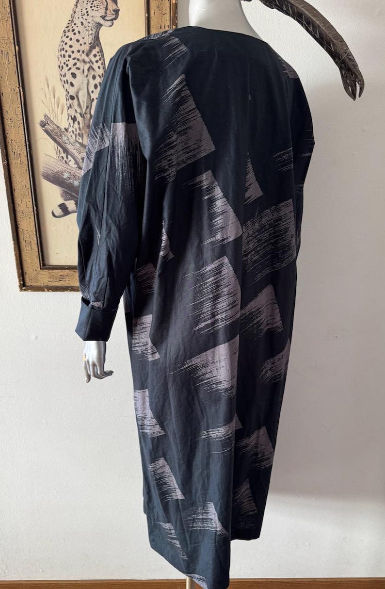 K&ouml;nnte beinhalten: Ein langes, schwarzes Kleid mit einem grauen Pinselstrichmuster. Das Kleid hat lange &Auml;rmel mit gekn&ouml;pften Manschetten. Das Kleidungsst&uuml;ck wird auf einer Schaufensterpuppe pr&auml;sentiert. Der Hintergrund umfasst ein gerahmtes Bild eines Leoparden und eine dekorative Feder.
