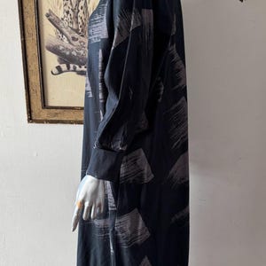 K&ouml;nnte beinhalten: Schwarzes Kleid mit langen &Auml;rmeln und V-Ausschnitt, mit einem Muster aus grauen Pinselstrichen. Das Kleid hat eine lockere Passform und lange &Auml;rmel mit gekn&ouml;pften Manschetten. Das Kleid wird auf einer Schaufensterpuppe pr&auml;sentiert.