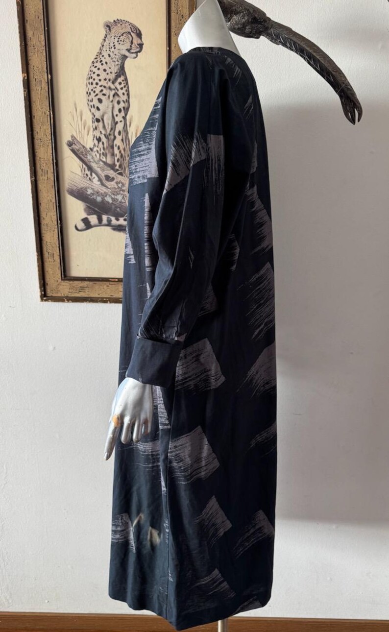 K&ouml;nnte beinhalten: Schwarzes Kleid mit langen &Auml;rmeln und V-Ausschnitt, mit einem Muster aus grauen Pinselstrichen. Das Kleid hat eine lockere Passform und lange &Auml;rmel mit gekn&ouml;pften Manschetten. Das Kleid wird auf einer Schaufensterpuppe pr&auml;sentiert.