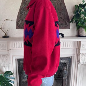 May include: A red jacket with a geometric pattern in black, purple, and red. The jacket has a ribbed hem and cuffs, a button-up front, and a collared neckline. The jacket is displayed on a mannequin.