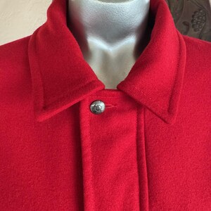 May include: Close-up of a red wool jacket with a black geometric pattern at the bottom. The jacket has a classic collar and a single silver button. The fabric appears to be a thick, warm material.