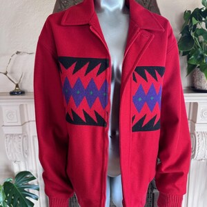 May include: A red zip-up jacket with a collar and ribbed cuffs. The jacket features a geometric pattern in black, purple, and green across the chest. The jacket is displayed on a mannequin.