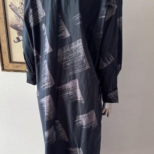 K&ouml;nnte beinhalten: Ein langes, schwarzes Kleid mit einem grauen Pinselstrichmuster. Das Kleid hat lange &Auml;rmel und eine lockere Passform. Der Stoff scheint leicht und flie&szlig;end zu sein. Das Kleid wird auf einer Schaufensterpuppe pr&auml;sentiert.