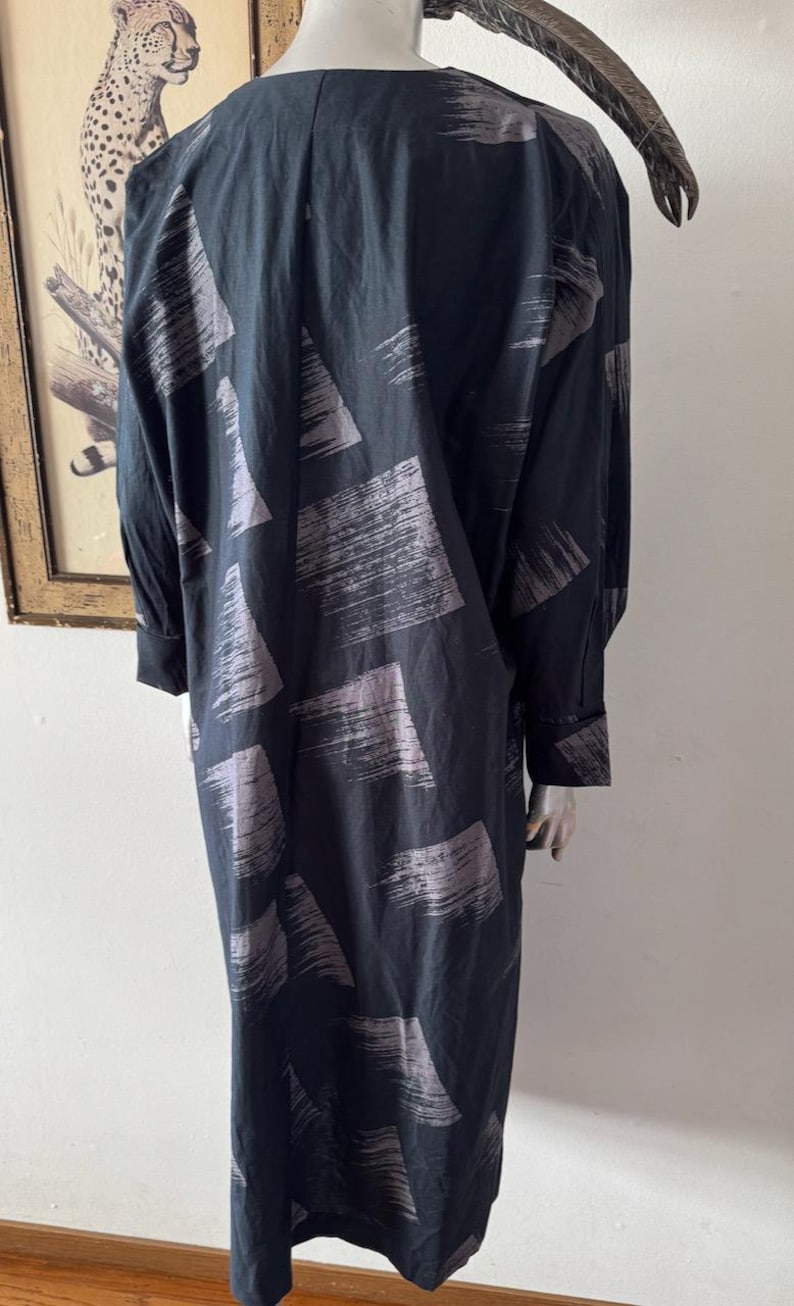 K&ouml;nnte beinhalten: Ein langes, schwarzes Kleid mit einem grauen Pinselstrichmuster. Das Kleid hat lange &Auml;rmel und eine lockere Passform. Der Stoff scheint leicht und flie&szlig;end zu sein. Das Kleid wird auf einer Schaufensterpuppe pr&auml;sentiert.