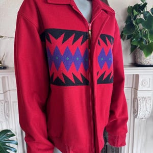 May include: A red zip-up jacket with a geometric pattern in black, purple, and green. The jacket has a collar and ribbed cuffs. The zipper is gold-colored.