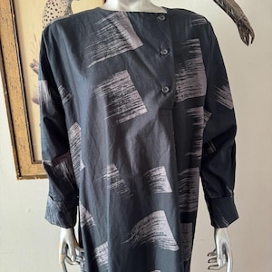 K&ouml;nnte beinhalten: Schwarzes Langarmkleid mit einem Pinselstrichmuster in Graut&ouml;nen. Das Kleid hat einen Knopfverschluss an der Schulter und an den Manschetten. Das Kleid wird auf einer Schaufensterpuppe pr&auml;sentiert.