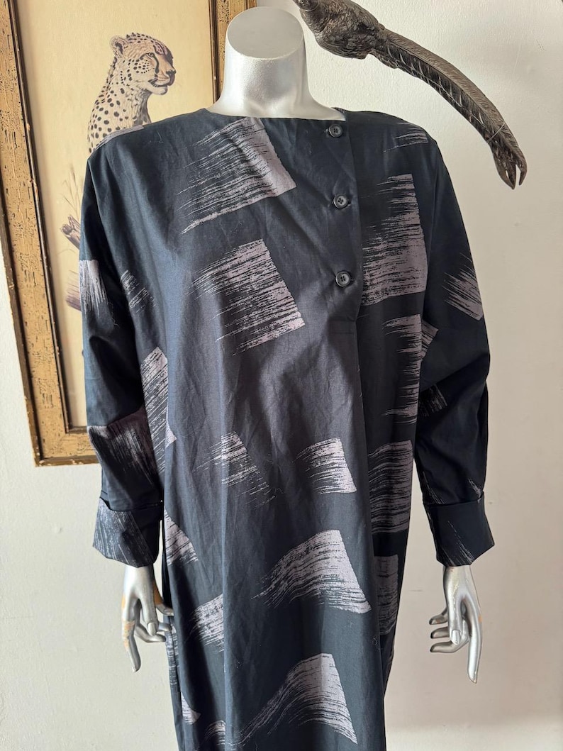 K&ouml;nnte beinhalten: Schwarzes Langarmkleid mit einem Pinselstrichmuster in Graut&ouml;nen. Das Kleid hat einen Knopfverschluss an der Schulter und an den Manschetten. Das Kleid wird auf einer Schaufensterpuppe pr&auml;sentiert.