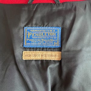 May include: Close-up of the inside of a black jacket with a red lining. The jacket features two rectangular tags. The top tag is blue with white text that reads "PENDLETON". The bottom tag is tan and reads "HIGH GRADE WESTERNWEAR".