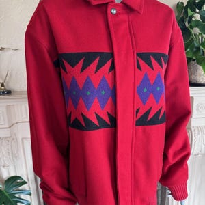 May include: A red jacket with a geometric pattern in black, purple, and green. The jacket has a collar, button closure, and ribbed cuffs. The design is a bold statement piece.