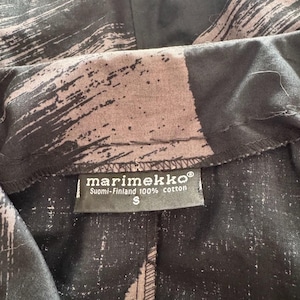 K&ouml;nnte beinhalten: Nahaufnahme eines schwarz-braunen Stoffes mit Pinselstrichmuster. Ein Etikett zeigt "marimekko" und "Suomi-Finland 100% cotton" mit der Gr&ouml;&szlig;e "S".
