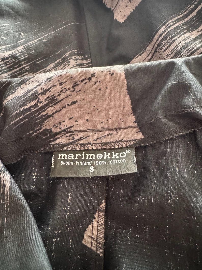 K&ouml;nnte beinhalten: Nahaufnahme eines schwarz-braunen Stoffes mit Pinselstrichmuster. Ein Etikett zeigt "marimekko" und "Suomi-Finland 100% cotton" mit der Gr&ouml;&szlig;e "S".