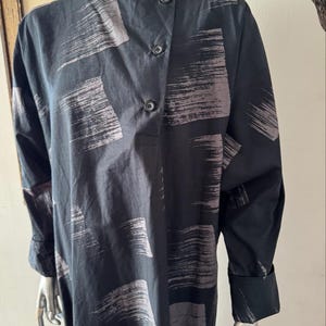 K&ouml;nnte beinhalten: Schwarzes Langarmkleid mit einem Pinselstrichmuster in Grau. Das Kleid hat einen Knopfverschluss und lange &Auml;rmel mit gekn&ouml;pften Manschetten. Das Kleid wird auf einer Schaufensterpuppe pr&auml;sentiert.