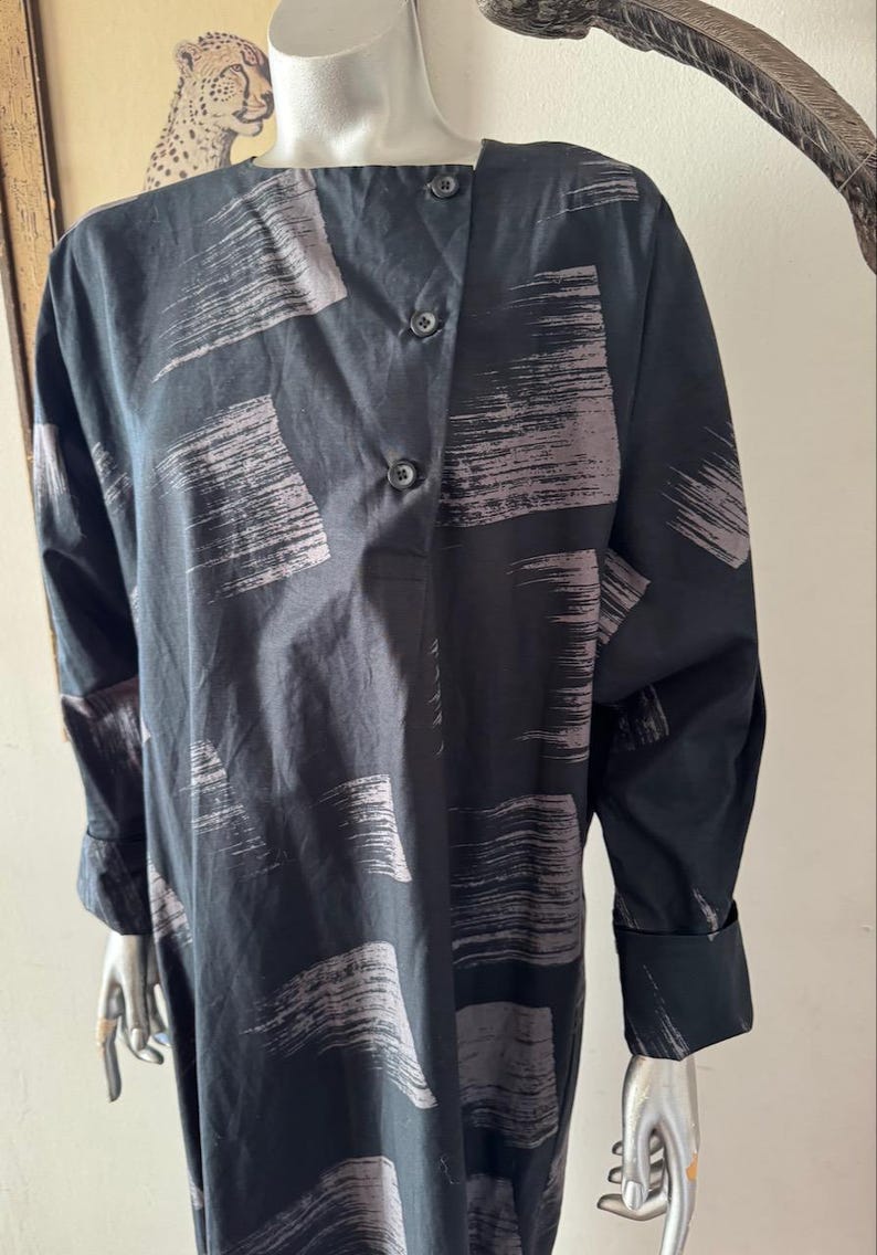 K&ouml;nnte beinhalten: Schwarzes Langarmkleid mit einem Pinselstrichmuster in Grau. Das Kleid hat einen Knopfverschluss und lange &Auml;rmel mit gekn&ouml;pften Manschetten. Das Kleid wird auf einer Schaufensterpuppe pr&auml;sentiert.
