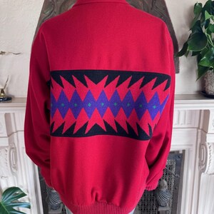 May include: A red zip-up sweater with a geometric pattern across the back. The pattern features black, purple, and green diamond shapes. The sweater has a high collar and ribbed cuffs and hem.