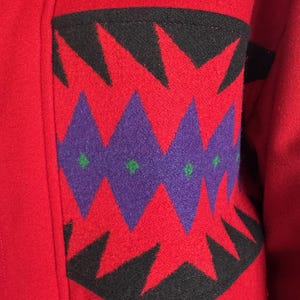 May include: Close-up of a red wool coat with a decorative pocket. The pocket features a geometric design with black, purple, and green accents. The coat's fabric appears thick and warm, suggesting a winter garment.
