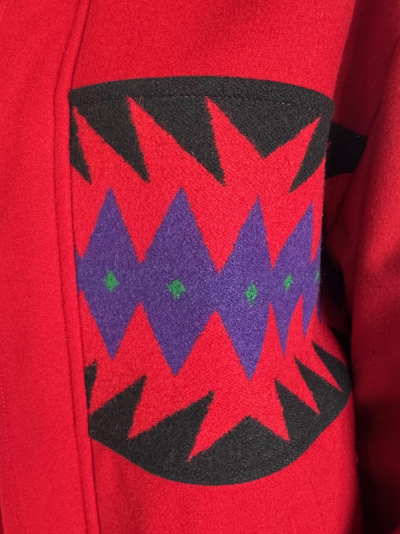 May include: Close-up of a red wool coat with a decorative pocket. The pocket features a geometric design with black, purple, and green accents. The coat's fabric appears thick and warm, suggesting a winter garment.