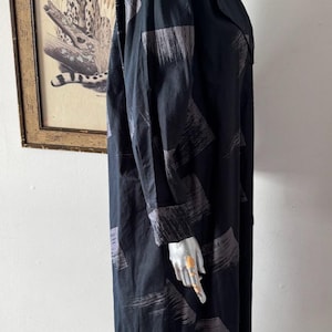 K&ouml;nnte beinhalten: Schwarzes langes Kleid mit grauem Pinselstrichmuster. Das Kleid hat lange &Auml;rmel und einen Reverskragen. Das Kleid wird auf einer Schaufensterpuppe pr&auml;sentiert. Im Hintergrund befindet sich ein gerahmtes Bild eines Leoparden.