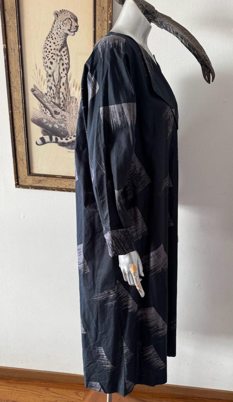 K&ouml;nnte beinhalten: Schwarzes langes Kleid mit grauem Pinselstrichmuster. Das Kleid hat lange &Auml;rmel und einen Reverskragen. Das Kleid wird auf einer Schaufensterpuppe pr&auml;sentiert. Im Hintergrund befindet sich ein gerahmtes Bild eines Leoparden.