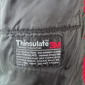 May include: Close-up of a black garment lining with a rectangular label. The label features the "Thinsulate" and "3M" logos, along with care instructions. The garment is red on the edges.