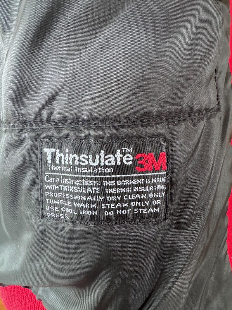 May include: Close-up of a black garment lining with a rectangular label. The label features the "Thinsulate" and "3M" logos, along with care instructions. The garment is red on the edges.