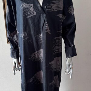 K&ouml;nnte beinhalten: Ein langes, dunkelblaues Kleid mit V-Ausschnitt und langen &Auml;rmeln. Das Kleid hat ein Muster aus grauen Pinselstrichen auf dem Stoff. Das Kleid ist am Ausschnitt gekn&ouml;pft und hat eine lockere Passform.