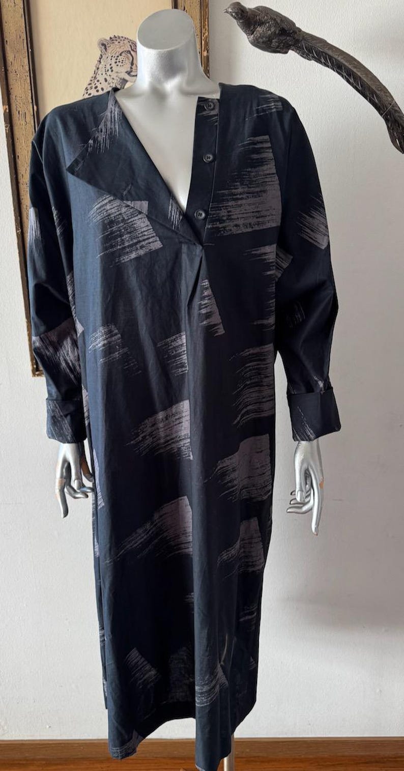 K&ouml;nnte beinhalten: Ein langes, dunkelblaues Kleid mit V-Ausschnitt und langen &Auml;rmeln. Das Kleid hat ein Muster aus grauen Pinselstrichen auf dem Stoff. Das Kleid ist am Ausschnitt gekn&ouml;pft und hat eine lockere Passform.