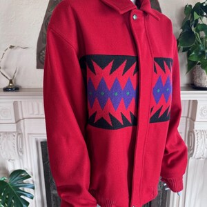May include: A red wool jacket with a black, purple, and green geometric pattern on the front. The jacket has a button closure, a collar, and ribbed cuffs. The jacket is displayed on a mannequin.
