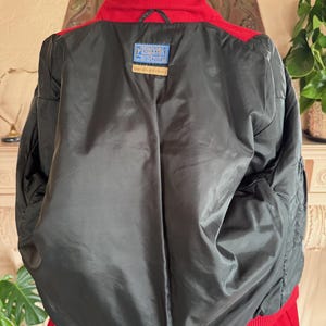 May include: A red and black bomber jacket with a red collar and cuffs. The jacket has a black lining and a label with text. The jacket is made of a wool blend.