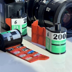 Puede incluir: Primer plano de tres cartuchos de película de 35 mm, dos son verdes y blancos con el texto "Fujicolor 200" y "6", el otro es negro con un patrón de cuadrícula. Un soporte para tarjetas SD rojo y naranja está en el fondo.