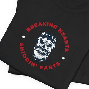May include: Black t-shirt with a white skull graphic wearing goggles and a beard. The text around the skull reads "Breaking Hearts Shiddin' Farts" in red.