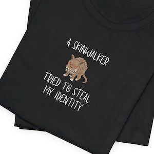 May include: Black t-shirt with a white graphic of a cartoon monster with red eyes and the text "A Skinwalker Tried To Steal My Identity".