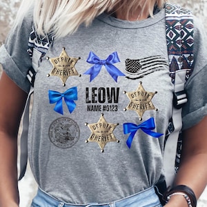 Peut inclure: Un t-shirt gris avec plusieurs badges "Deputy Sheriff" en forme d'étoile dorée, des nœuds bleus et un drapeau américain vieilli. Le nom "LEOW" et le numéro "0123" sont imprimés sous les nœuds.