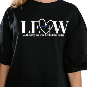 Peut inclure: T-shirt noir avec texte blanc et motif cœur. Le texte indique "LE♡W" avec une ligne bleue dans le cœur et une croix. En dessous, la phrase "...the praying wife behind the badge."