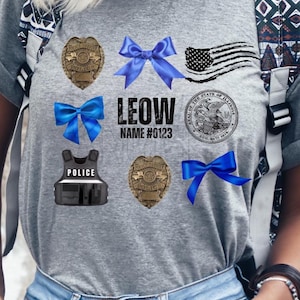 Peut inclure: T-shirt gris avec des graphiques sur le thème de la police. Comprend un badge de police, des nœuds bleus, un drapeau américain et le sceau de l'État de l'Illinois. Le texte "LEOW NAME #0123" est également affiché. Un gilet pare-balles noir est également présent.