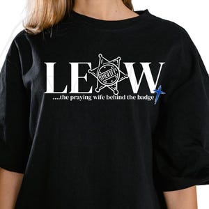Peut inclure: T-shirt noir avec texte blanc et un insigne de shérif. Le texte "LEW" est surmonté d'un insigne en forme d'étoile avec le mot "SHERIFF" à l'intérieur. En dessous, on lit "...the praying wife behind the badge" et une croix bleue.