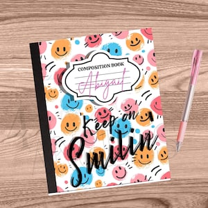 Puede incluir: Cuaderno de composición con un fondo blanco cubierto de caras sonrientes de colores. El cuaderno tiene el texto "Keep on Smilin" y "Abigail" en la portada, junto con un bolígrafo rosa.