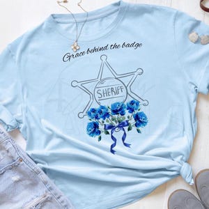 Peut inclure: T-shirt bleu clair avec un motif d'insigne de shérif, des roses bleues et le texte "Grace behind the badge". Le motif est centré sur le devant. Le t-shirt est en matière souple. Vêtement décontracté.