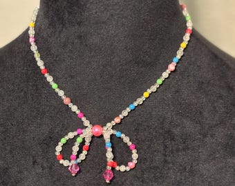 May include: A colourful beaded necklace with a bow-shaped pendant. The necklace features small, round beads in white, pink, red, yellow, green, and blue. The bow pendant has a large pink bead in the centre and two pink crystal-like drops.