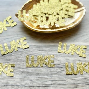 May include: A collection of gold glitter die-cut paper pieces, each spelling the name LUKE in a bold sans-serif font. Several individual pieces are scattered around a gold-colored leaf-shaped dish that also contains a pile of the same cutouts.