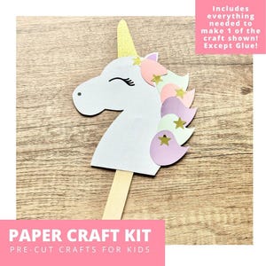 May include: A paper craft kit featuring a white unicorn head cutout with a gold glitter horn, a pastel-colored mane, and small gold star accents. The unicorn is attached to a wooden stick. Text reads: Includes everything needed to make 1 of the craft shown! Except Glue! PAPER CRAFT KIT PRE-CUT CRAFTS FOR KIDS