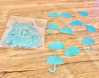 May include: A collection of light blue glittery paper cutouts shaped like umbrellas, clouds, and raindrops. Some of these shapes are contained within a translucent white plastic bag featuring a subtle white polka dot pattern.