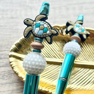 May include: Two teal and gold pens with decorative elements. Each pen features a white beaded sphere, a brown bead, and a turtle-shaped charm with teal and tan accents. The pens rest on a gold leaf-shaped tray.