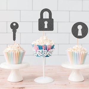 May include: Three cupcakes with white frosting and decorative toppers. One cupcake has a key topper, another a padlock, and the third a keyhole. The cupcakes sit on white cake stands and a decorative stand, with a pastel-colored liner.