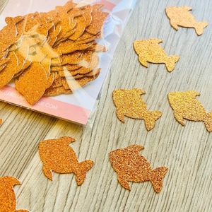 May include: A clear plastic bag containing orange glittery shark-shaped confetti pieces, with several identical orange glittery shark-shaped confetti pieces scattered outside the bag. The bag features a label with the text Little Bella Boutique.