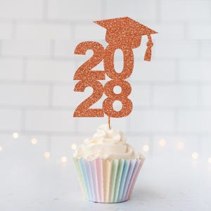 May include: A copper-colored glitter graduation cupcake topper featuring the numbers 2028 topped with a mortarboard cap. The topper is inserted into a white frosted cupcake with a pastel-colored pleated paper liner, creating a festive display.