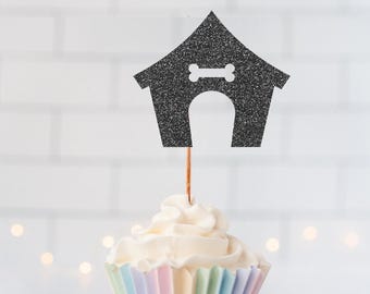 May include: A cupcake topper shaped like a black glittery dog house with a white bone cutout above the arched entrance, attached to a thin wooden pick and inserted into a cupcake with a pastel-colored paper liner featuring vertical pleats.