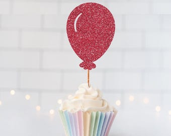 May include: A red glitter balloon cupcake topper featuring a shiny, textured surface and a white curved highlight line, attached to a thin wooden pick and inserted into a white frosted cupcake with a pastel striped paper liner.
