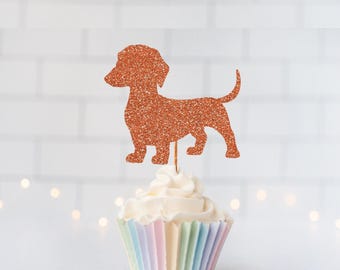 May include: A cupcake topper featuring a glittery copper-colored silhouette of a dachshund dog. The cutout is attached to a thin wooden pick and is inserted into a cupcake with white frosting and a pastel-colored pleated paper liner.