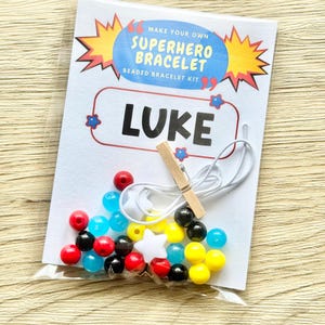 May include: A DIY superhero bracelet kit in a clear plastic bag with a white card featuring the text MAKE YOUR OWN SUPERHERO BRACELET BEADED BRACELET KIT and the name LUKE. The kit includes colorful red, blue, yellow, and black beads, white star-shaped beads, a white elastic cord, and a small wooden clothespin.
